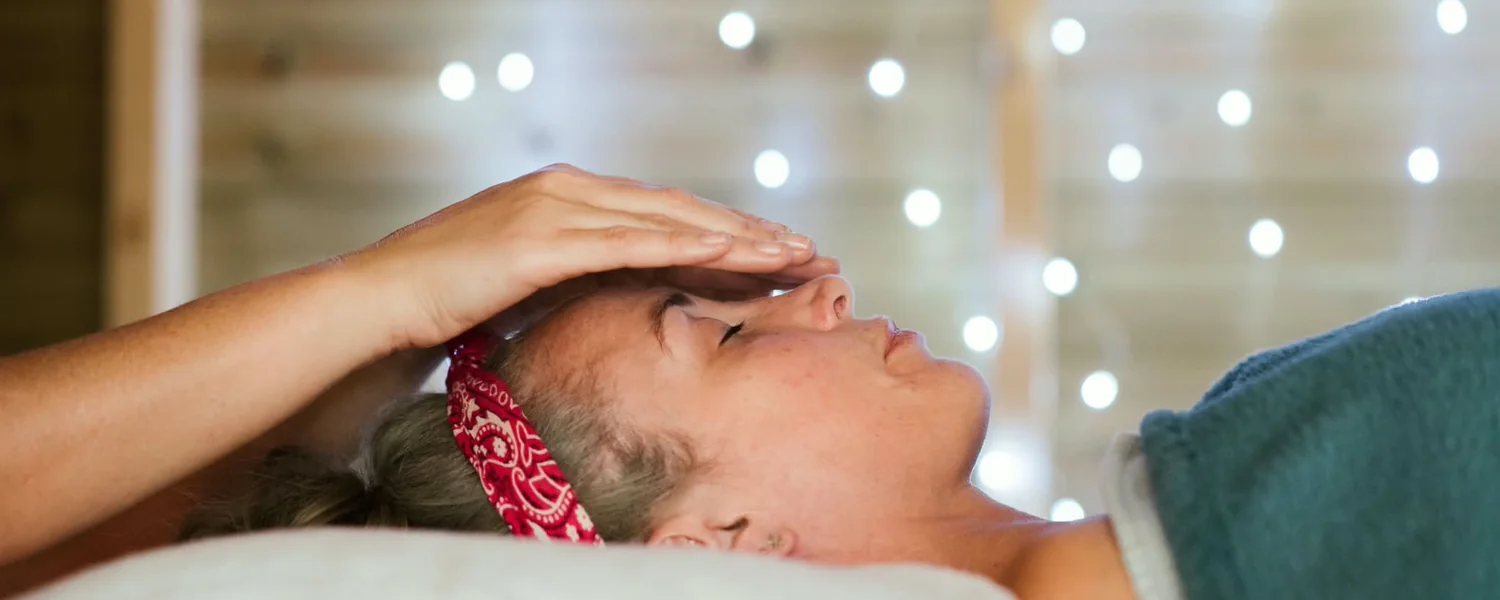 A practitioner's hands resting above a client's forehead, warm light behind